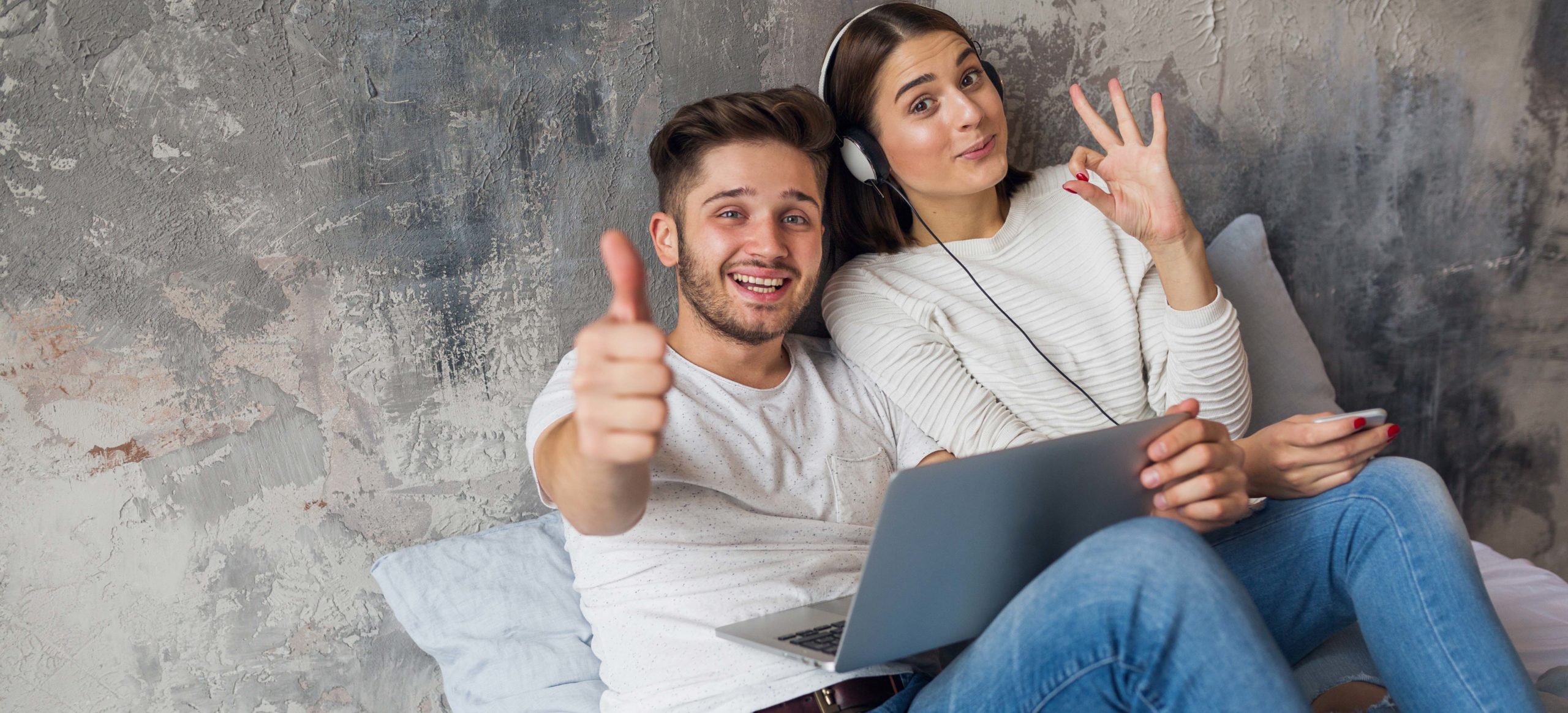 young smiling couple sitting on bed at home in casual outfit, man working freelance on laptop, woman listening to music on headphones, spending happy time together, positive emotion, looking in camera